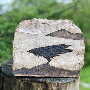 May include: A ceramic tile featuring a black silhouette of a crow against a white background. The crow is perched on a small hill with a mountain range in the background.