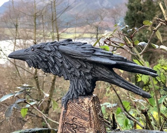 May include: A black ceramic raven sculpture with detailed feathers perched on a textured brown ceramic base. The sculpture is displayed on a tree stump outdoors.