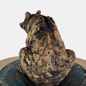 Op de afbeelding: Een keramische berensculptuur, van achteren gezien, zittend op een blauw en grijs oppervlak. De beer heeft een getextureerde, ruwe afwerking in donkerbruin en beige, die de details van de klei benadrukt. De oren van de beer zijn zichtbaar.