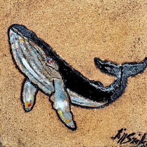 May include: A handcrafted whale art piece with a textured surface. The whale is depicted in black, white, and gray, with colorful accents. The artwork is set against a tan background. The artist's signature is visible in the bottom right corner.