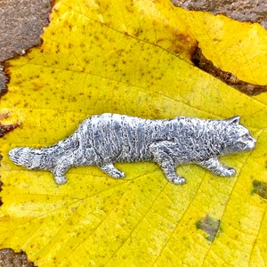 May include: A silver-colored metal sculpture of a cat lying on a yellow leaf. The cat is detailed with fur and has a realistic appearance.