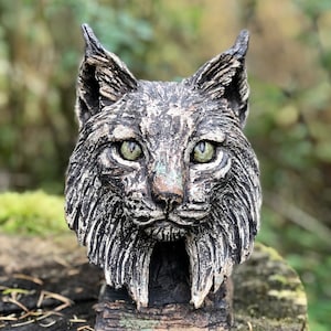 May include: A ceramic sculpture of a lynx head with a textured, realistic finish. The lynx has green eyes and a detailed, sculpted face.