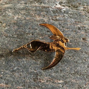 Puede incluir: Un broche de metal dorado que representa una liebre estilizada saltando sobre una luna creciente.