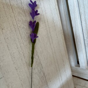 May include: A single purple felt lavender flower stem with green felt leaves on a white wooden background.