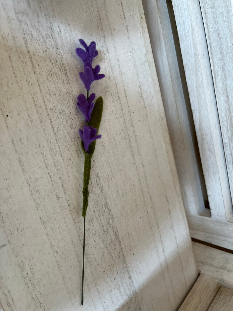 May include: A single purple felt lavender flower stem with green felt leaves on a white wooden background.