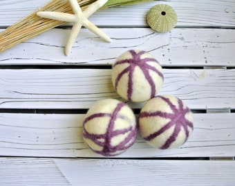 May include: Three white wool dryer balls with purple stripes. The balls are on a white wooden surface.