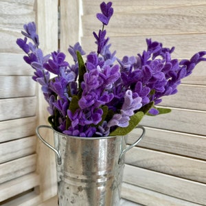 May include: A bouquet of purple lavender flowers in a galvanized metal vase with handles.