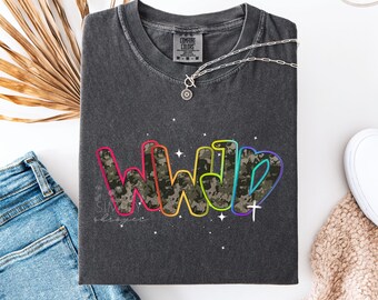 May include: A dark gray t-shirt with the letters "WWJD" in a colorful, outlined font with a camouflage pattern. A silver necklace and a pair of blue jeans are visible. The shirt is folded and has the Comfort Colors brand tag.