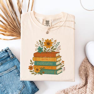 May include: Cream-colored Comfort Colors t-shirt featuring a stack of books with floral accents in orange, teal, and yellow. The design includes a necklace and is paired with denim jeans and a white shoe.