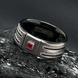 May include: A black titanium ring with a red gemstone and a cable-like design. The ring is engraved with the word "TITANIUM".