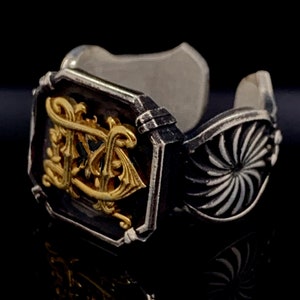 Op de afbeelding: Een zilveren ring met een goudkleurig ontwerp in het midden. De ring heeft een getextureerd, wervelend ontwerp aan de zijkant.