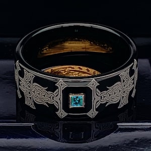 Könnte beinhalten: Ein schwarzer Keramikring mit einem silbernen keltischen Kreuzdesign und einem blauen Edelstein in der Mitte.