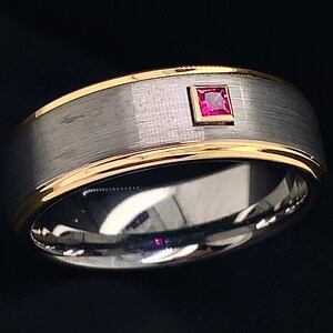 Puede incluir: Una banda de boda de plata y oro con una pequeña piedra preciosa roja en el centro.