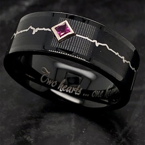 May include: Black tungsten carbide ring with a silver engraved heartbeat design and a small pink gemstone. The inscription "Two hearts... one heart" is engraved on the inside of the band.