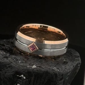 May include: A silver and rose gold wedding band with a single, square-shaped ruby gemstone.