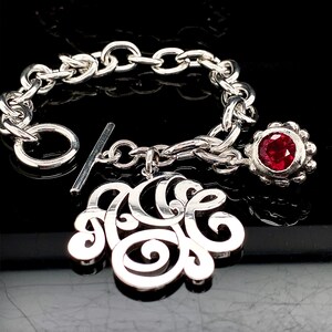Può includere: Un braccialetto d'argento con chiusura a scorrimento e un ciondolo con monogramma. Il braccialetto presenta anche un piccolo ciondolo con pietra preziosa rossa.