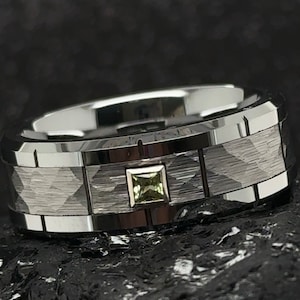 Puede incluir: Un anillo de carburo de tungsteno negro con acabado cepillado y una sola piedra preciosa verde de forma cuadrada engastada en el centro.