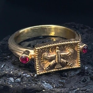 May include: A gold ring with a rectangular design featuring a stylized bird with wings outstretched. The ring is adorned with two small red gemstones.