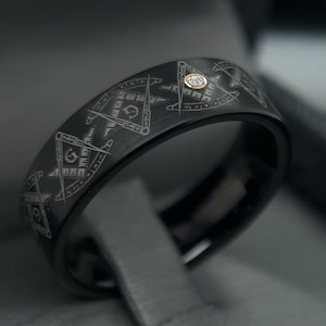 May include: A black tungsten carbide ring with a Masonic symbol design and a small diamond set in the center.