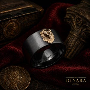 Könnte beinhalten: Ein breiter Ring aus gebürstetem schwarzem Metall mit einem goldfarbenen Löwenkopf-Emblem und kleinen eingesetzten Steinen. Die Innenseite des Rings ist mit dem Text Love you forever graviert. Das Design kombiniert ein dunkles, mattes Finish.