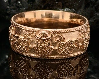 May include: A gold-colored ring with detailed relief carvings. The ring features a central face design with a beard, surrounded by ornate patterns and figures. The ring's design is reflected in the surface below.