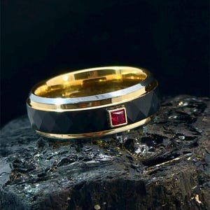 May include: A gold-toned ring with a black band and a square, red gemstone. The ring has a faceted design on the black band and a silver-colored edge. The ring is displayed on a dark, textured surface.