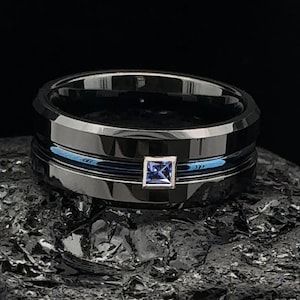 May include: A black tungsten ring featuring a blue sapphire gemstone. The ring has a polished finish with a blue inlay. The ring is displayed on a dark, textured surface.