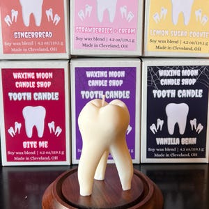 May include: A tooth-shaped candle made of soy wax, standing on a wooden base, with a variety of scented candle boxes in the background. The boxes are labeled with flavors like "Gingerbread" and "Vanilla Bean". Each candle is 4.2 oz / 119.1 g.