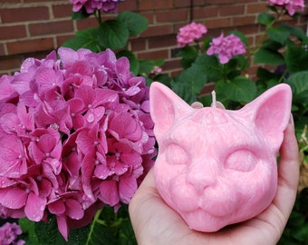 Puede incluir: Vela rosa con forma de gato con una cara detallada y los ojos cerrados. La vela se sostiene en una mano sobre un fondo de hortensias rosas.