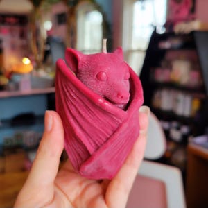 Peut inclure: Une bougie en forme de chauve-souris de couleur bordeaux, avec des ailes détaillées et une petite mèche. La bougie est tenue dans une main, mettant en valeur son design unique. Le visage de la chauve-souris est visible.