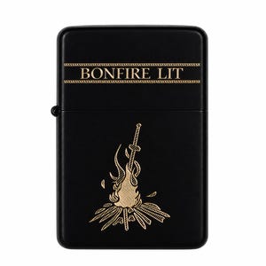 May include: A matte black metal flip-top lighter featuring a gold-coloured engraved design. The upper portion displays the text "BONFIRE LIT" between two horizontal lines. Below, a sword is depicted standing upright in a burning bonfire.