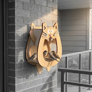Puede incluir: Un comedero para pájaros de madera con forma de gato, montado en una pared de ladrillo. El comedero tiene una gran abertura para que entren los pájaros y una bandeja para alpiste. Varios pájaros pequeños están posados en la bandeja, alimentándose. La cara del gato está detallada con ojos, bigotes y una nariz rosa.