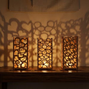May include: Three wooden candle holders with detailed cut-out patterns, projecting heart-shaped shadows. Each holder contains a lit candle, creating a warm glow. The holders are rectangular and sit on a dark wooden surface.
