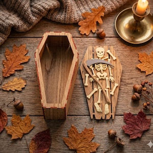 May include: Two wooden coffin-shaped boxes, one open and one with a skeleton and scythe design, sit on a wooden surface. Autumn leaves, acorns, and a lit candle in a brass holder surround the boxes, creating a Halloween theme.