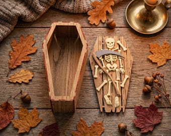 May include: Two wooden coffin-shaped boxes, one open and one with a skeleton and scythe design, sit on a wooden surface. Autumn leaves, acorns, and a lit candle in a brass holder surround the boxes, creating a Halloween theme.