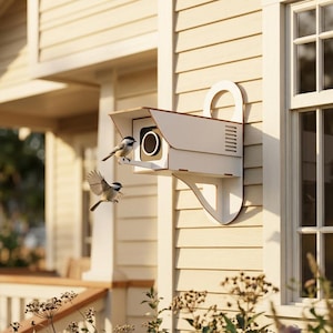 May include: A cream-colored birdhouse designed to resemble a security camera, mounted on a light-colored exterior wall. Two small birds are perched on and near the birdhouse. The birdhouse has a curved handle and a lens-like opening.