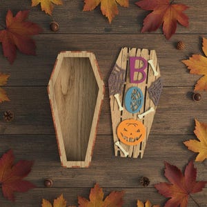 May include: Wooden coffin-shaped box and a decorative coffin with the word "BOO" spelled out with purple, blue, and orange accents. The decorative coffin has a carved pumpkin and bone accents. Autumn leaves and acorns surround the coffins.