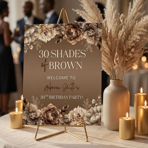 Puede incluir: Un letrero rectangular sobre un caballete de metal dorado con ilustraciones florales marrones y el texto 30 SHADES of BROWN WELCOME TO Aubreon Smith's 30TH BIRTHDAY PARTY. El letrero se muestra con velas doradas y un jarr&oacute;n con hierba.