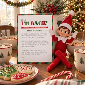 May include: A white rectangular sign with red and green text reading Hey! I'M BACK! AND READY TO MAKE NEW MEMORIES WITH ALLIE & THOMAS, alongside a plush elf doll wearing a red suit and hat with white trim, positioned next to holiday treats.