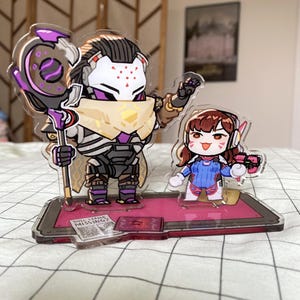 May include: Acrylic figurines of two characters from the video game Overwatch. One figure is a tall, armored character holding a staff. The other is a smaller character in a blue and white outfit, holding a pink gun. The base has the text "NULL LEADER MISSING?"