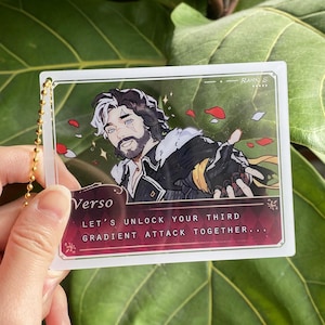 Op de afbeelding: Een heldere acryl bedel met een gouden ketting. De bedel toont een mannelijk anime-personage met zwart en wit haar, een baard en een zwart-gouden outfit. De tekst "Verso" en "Let's unlock your third gradient attack together..." zijn zichtbaar.