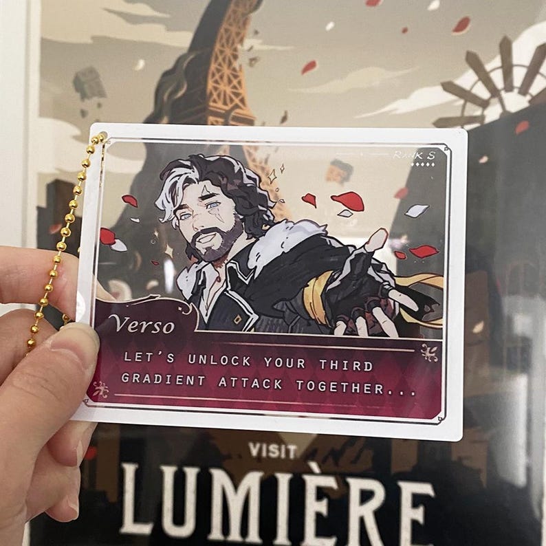 Op de afbeelding: Een heldere acryl bedel met een illustratie van een personage genaamd Verso. Het kunstwerk toont een man met wit en donker haar, in een zwart en gouden outfit. De bedel bevat de tekst "Let's unlock your third gradient attack together..."