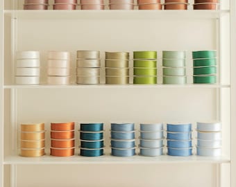 May include: A shelf with multiple spools of ribbon in various colors, including white, pink, green, blue, and orange. The spools are arranged in rows on the shelf.