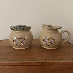 May include: A tan ceramic creamer and sugar bowl set. The sugar bowl has a lid. Both pieces are decorated with a heart-shaped floral design in pink, blue, and white. The set is displayed on a wooden surface.