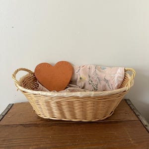 Può includere: Un cesto di vimini con un sottopentola in terracotta a forma di cuore al suo interno. Il sottopentola ha le parole "Ready, There, Near" e "Walk Your Own Path". Il cesto è su una superficie di legno.