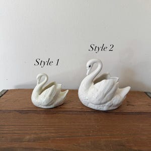 May include: Two white ceramic swan ornaments, labelled "Style 1" and "Style 2". The swans are displayed on a wooden surface against a neutral background. The figurines have detailed feather textures and curved necks.
