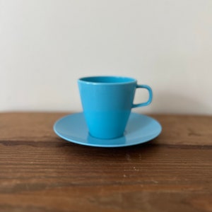 May include: A light blue ceramic coffee cup and saucer set. The cup has a rounded shape and a handle. The saucer is round and has a slightly raised edge.