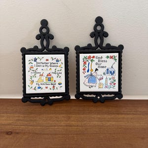 Puede incluir: Dos placas de pared rectangulares con marcos de metal negro y azulejos de cerámica blanca. El azulejo izquierdo muestra una escena de cocina con el texto: No Matter Where I Serve My Guests It Seems They Like My Kitchen Best. El azulejo derecho muestra a una persona cocinando con el texto: God Bless Our Home.