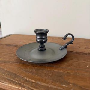 May include: A small, antique-style pewter candlestick holder. The holder has a circular base with a curved handle and a central cup for a candle. The metal has a dark, matte finish, giving it a vintage appearance. The candlestick holder is designed to sit on a flat surface.