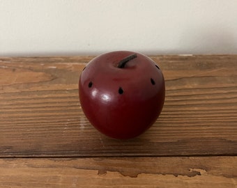 May include: A red apple-shaped ceramic incense burner with small holes on the surface. The burner is sitting on a wooden surface.
